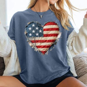 May include: A blue t-shirt featuring a heart-shaped American flag design. The flag is made of red and white stripes with a blue section containing white stars. The design has a distressed, vintage look, with frayed edges.