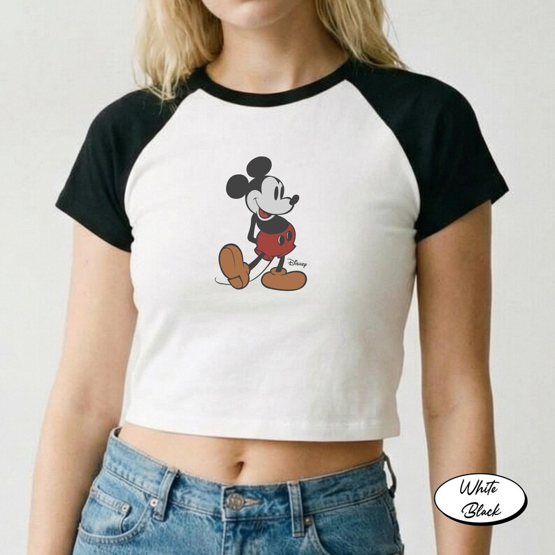 May include: White and black raglan crop top featuring a Mickey Mouse graphic. The graphic shows Mickey Mouse in his classic outfit. The shirt has black sleeves and a white body. The Disney logo is visible on the bottom right of the graphic.