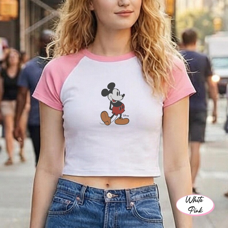 May include: White and pink raglan crop top featuring a Mickey Mouse graphic. The shirt has a white body with pink sleeves and neckline. The Mickey Mouse graphic is in the center of the shirt. The shirt is paired with blue jeans.