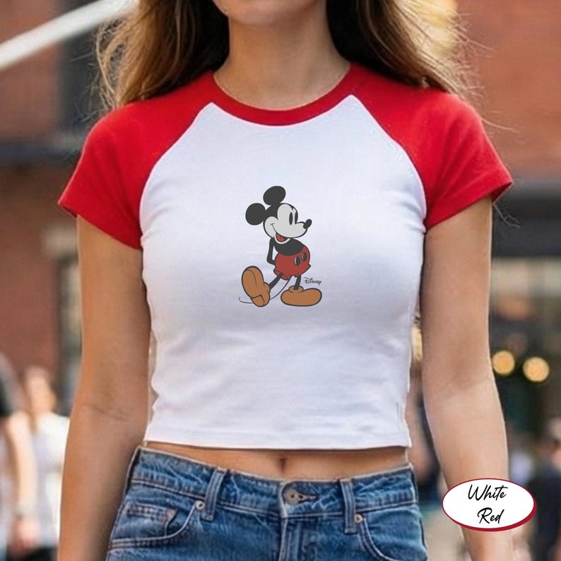 May include: White crop top with red short sleeves featuring a Mickey Mouse graphic. The cartoon character is depicted in a walking pose, wearing red shorts and yellow shoes. The shirt is a casual style.