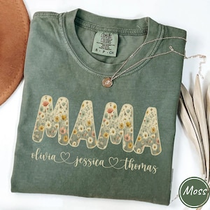 May include: Moss green t-shirt with the word "MAMA" in a floral design. Below the word "MAMA" are the names "olivia", "jessica", and "thomas". A silver necklace with a pendant rests on the shirt. The shirt is labeled "Comfort Colors".