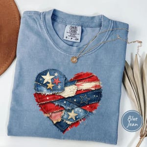 May include: A blue jean-colored t-shirt with a heart-shaped design made of denim patches in red, white, and blue, with star accents. The shirt has a crew neck and short sleeves. A gold necklace with a pendant is draped across the shirt.