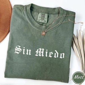 May include: An olive green t-shirt with the words "Sin Miedo" in white gothic lettering. A gold-colored necklace with a pendant rests on the shirt. The shirt is folded and has a Comfort Colors tag. The color of the shirt is labeled "Moss".
