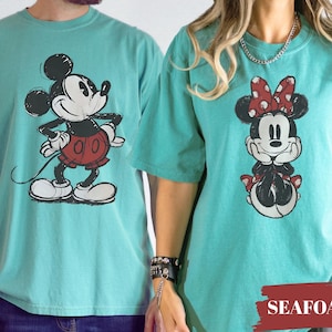 May include: Seafoam green t-shirts featuring Mickey and Minnie Mouse. Mickey is in a red shorts and Minnie is wearing a red polka dot bow. The shirts are a comfortable, casual style.