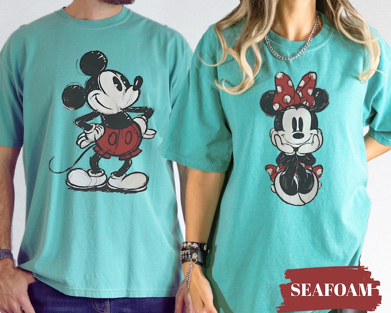 May include: Seafoam green t-shirts featuring Mickey and Minnie Mouse. Mickey is in a red shorts and Minnie is wearing a red polka dot bow. The shirts are a comfortable, casual style.