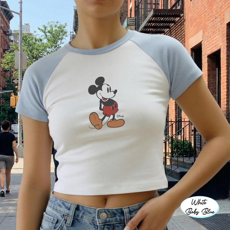 May include: White and baby blue raglan crop top featuring a Mickey Mouse graphic. The shirt has a white body with baby blue sleeves and neckline. The Mickey Mouse illustration is in classic colors.