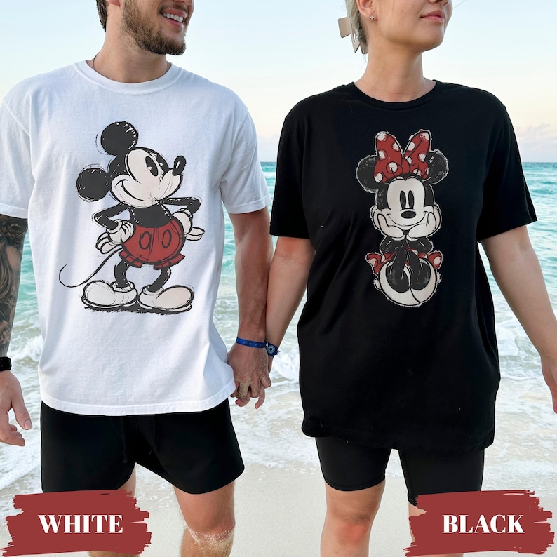 May include: Two people wearing matching graphic t-shirts on a beach. One shirt is white with a Mickey Mouse design, and the other is black with a Minnie Mouse design. Both shirts have the character's image in a vintage style.