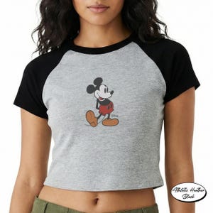 May include: Gray raglan crop top featuring a classic Mickey Mouse graphic. The shirt has black sleeves and a round neckline. The graphic shows Mickey Mouse in his signature red shorts and yellow shoes. The shirt is labeled "Athletic Heather Black".