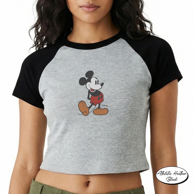 May include: Gray raglan crop top featuring a classic Mickey Mouse graphic. The shirt has black sleeves and a round neckline. The graphic shows Mickey Mouse in his signature red shorts and yellow shoes. The shirt is labeled "Athletic Heather Black".
