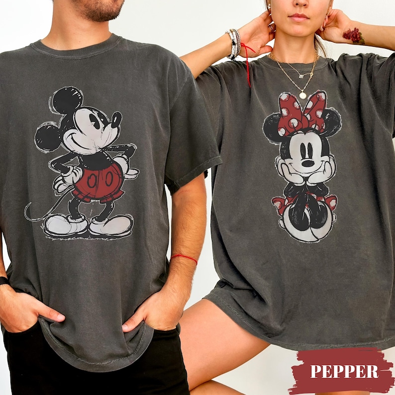 May include: Two charcoal gray t-shirts featuring Mickey and Minnie Mouse. Mickey is depicted in a red shorts and white gloves, while Minnie wears a red bow. The shirts have a vintage, distressed look.