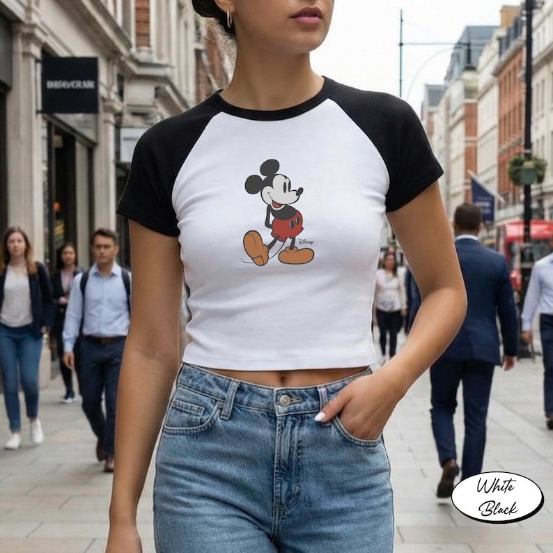 May include: White crop top with black sleeves featuring a Mickey Mouse graphic. The shirt is a baseball style tee. The image also shows a person wearing blue jeans. The shirt has the words "White" and "Black" on the bottom right.