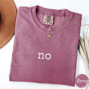 May include: A berry-colored t-shirt with the word "no" printed in white. The shirt has a crew neck and short sleeves. A gold-colored necklace with a pendant is draped across the shirt. The shirt is folded and placed on a white surface.