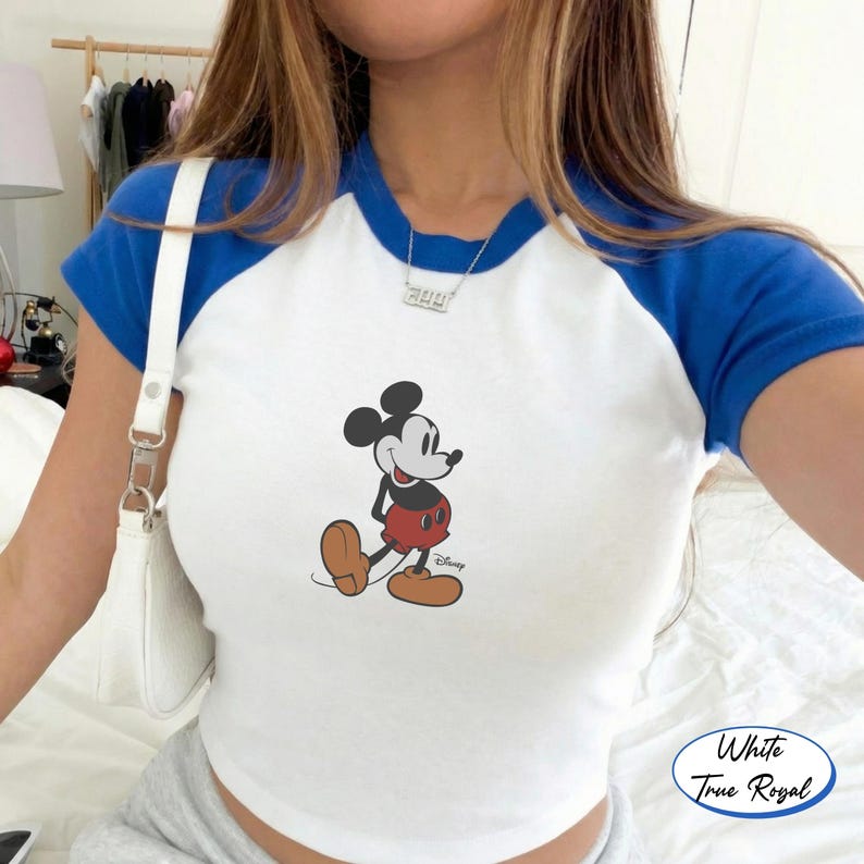 May include: White and royal blue raglan style crop top featuring a Mickey Mouse graphic. The shirt has a white body with royal blue sleeves and neckline. The Mickey Mouse graphic is in the center of the shirt.