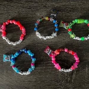 May include: Five colorful beaded bracelets with candy wrappers attached. The bracelets feature various colors like pink, red, blue, and green, with white letter beads spelling out words like "Twizzlers" and "Nerds".