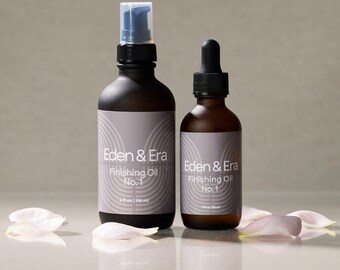 May include: Two dark brown glass bottles of Eden & Era Finishing Oil No. 1. One bottle has a spray pump, the other a dropper. Each bottle has a gray label with white text. The bottles are surrounded by white and pink flower petals.