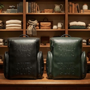 May include: Two rectangular backpacks, one black and one dark green, featuring the embossed text BANTU ANTS TRAVEL CLUB on the front. Each bag has a top handle, two side pockets, and a structured design, displayed side by side on a wooden surface.