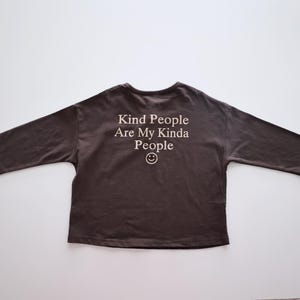 May include: A dark brown, long-sleeve shirt with the text "Kind People Are My Kinda People" and a smiley face graphic. The shirt is laid flat on a white surface, showcasing its design.