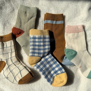 May include: A collection of five pairs of ankle socks featuring various patterns and colors, including a cream pair with a blue grid, a sage green pair with a rust heel, a yellow and blue plaid pair, a solid rust pair, and a cream pair with green accents.