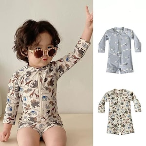 May include: A child wearing a beige and brown animal-print swimsuit with a front zipper and long sleeves. The swimsuit features illustrations of elephants, giraffes, and other animals. Two additional swimsuits are shown, one light blue with clouds and one with the same animal print.