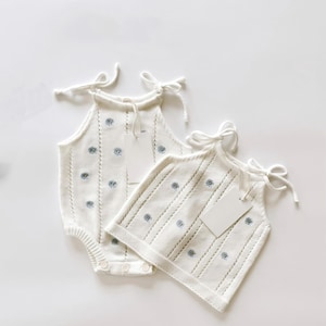 Könnte beinhalten: Zwei weiße gestrickte Baby-Outfits. Eines ist ein Strampler mit Druckknöpfen, das andere ein Tanktop. Beide haben blaue Blumenstickereien und Träger zum Binden. Jedes Kleidungsstück hat ein leeres Etikett. Die Outfits werden auf weißem Hintergrund präsentiert.