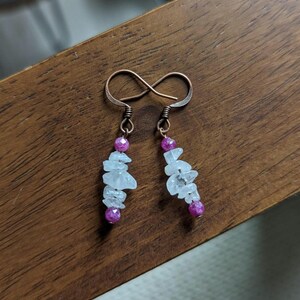 May include: A pair of handmade earrings with copper-colored ear wires. Each earring features a strand of light blue, irregularly shaped stones and two small, round, magenta-colored beads. The earrings are displayed on a wooden surface.