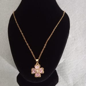 Könnte beinhalten: Eine goldfarbene Halskette mit einem vierblättrigen Kleeblatt-Anhänger. Der Anhänger ist mit rosa Edelsteinen und einem zentralen klaren Stein besetzt. Die Halskette wird auf einer schwarzen Schmuckbüste präsentiert.