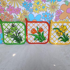 May include: Three square pot holders with floral designs. The pot holders have a white background with a grid pattern and are trimmed in green, orange, and yellow. Each features a different flower: green irises, orange tulips, and yellow daffodils.