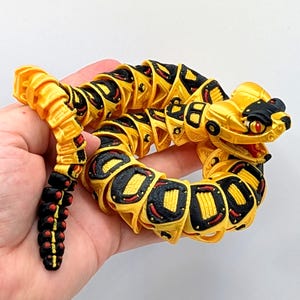May include: A yellow, black, and red articulated snake toy. The toy is held in a hand and has a segmented body with detailed features. The head has red eyes and black accents. The tail is black with red dots.