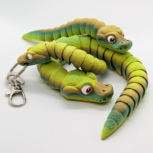May include: Three articulated toy snakes in shades of green, brown, and yellow are arranged on a white surface. One snake has a keychain attachment. The snakes have red eyes and are segmented to allow for movement.
