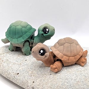 May include: Three 3D-printed turtle figurines in shades of green and brown rest on a textured grey surface. The turtles have large, expressive eyes and detailed shell patterns. The figurines are designed with a whimsical, cartoonish aesthetic.