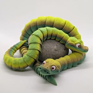 May include: A 3D-printed articulated snake toy in shades of green and yellow, coiled around a grey stone. The snake has a detailed, segmented body and cartoonish features, including red eyes.