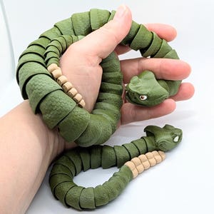 May include: A green articulated snake toy with a textured surface and beige accents. The snake is held in a hand, demonstrating its flexible design and detailed head with small eyes. The toy is designed to be bendable.