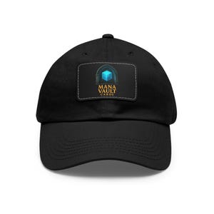 May include: Black baseball cap with a curved brim. The cap features a rectangular patch with the text "MANA VAULT CARDS" and a graphic of a blue cube inside an archway.