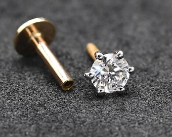 May include: Two pieces of jewelry: a gold-colored flat-backed stud and a silver-colored stud with a large, clear, round gemstone. The gemstone is faceted and reflects light. The jewelry is set against a dark, textured background.