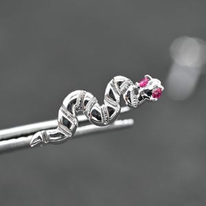 May include: A silver snake-shaped jewellery piece with a textured body and red gemstone eyes. The serpent is held by tweezers against a blurred grey background. The jewellery piece is a unique accessory.