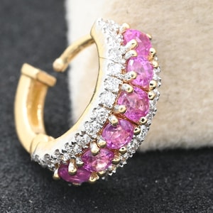 May include: A gold-toned hoop earring with a row of oval, pink gemstones and smaller, clear gemstones. The earring is set against a dark surface with a neutral background.