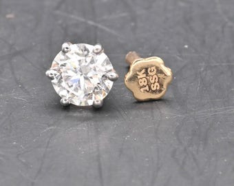 May include: A sparkling, round-cut diamond stud earring with a six-prong setting. Beside it, a gold earring back with the markings "18K SSG". The jewelry is set against a dark gray background.