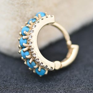 May include: A gold-colored hoop earring with a row of small, bright turquoise stones and clear accent stones. The earring is set against a dark background, highlighting the jewelry's details.