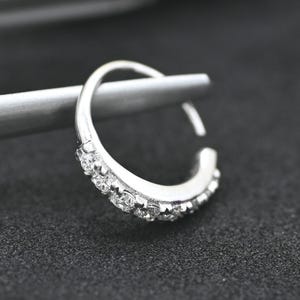 May include: A silver-colored nose ring with a row of small, clear gemstones. The ring is a hoop design, with the gemstones set along the outer curve. The jewelry is shown against a dark background.