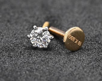May include: A close-up of a diamond stud earring with a six-prong setting. The earring features a brilliant-cut diamond and a gold post. The back of the earring is stamped with "18K SSG".