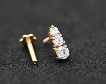 May include: A gold-colored earring stud with three clear, round gemstones in a vertical row. The earring post and the decorative backing are also gold. The background is a dark, textured surface.