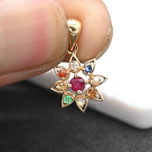 May include: A gold pendant in the shape of a flower, featuring a central ruby surrounded by various gemstones. The pendant is held up by a finger and thumb.