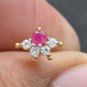 Pode incluir: Um brinco de pressão dourado em forma de estrela com uma pedra preciosa rosa redonda central e cinco pedras preciosas redondas e transparentes circundantes. O brinco está sobre um fundo desfocado.