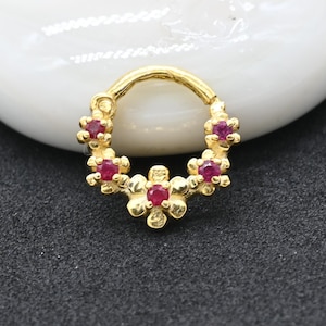 May include: A gold hoop ring featuring a series of five small flower-shaped metal settings, each centered with a tiny round red gemstone. The circular band is crafted from a polished gold-toned metal, creating a decorative floral arrangement.