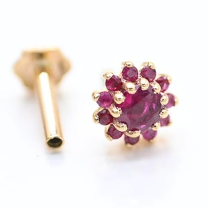 Puede incluir: Un pendiente de botón dorado con un diseño floral. El centro es una gema grande, redonda y de color rojo intenso, rodeada de gemas rojas más pequeñas a juego. El poste del pendiente también es de color dorado.