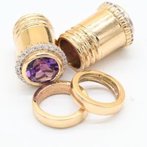May include: Gold-toned jewelry set featuring two cylindrical pendants with diamond accents and a central amethyst stone. Two gold rings are also present. The jewelry is displayed on a white surface.
