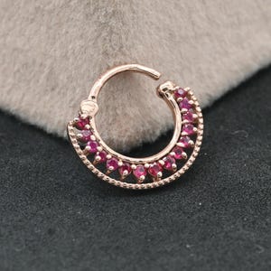 Peut inclure: Un anneau en or rose doté d'un mécanisme à charnière. Le bord extérieur est orné d'une rangée de petites pierres précieuses rondes d'un rose vif, serties dans une monture en métal perlée, formant un bijou circulaire pour le corps.
