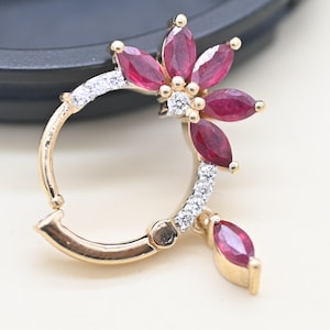Puede incluir: Pendiente dorado con diseño floral. La flor presenta seis gemas rojas oscuras con corte marquesa y pequeñas gemas redondas y claras. Una sola gema roja cuelga debajo. El pendiente tiene un cierre de bisagra.