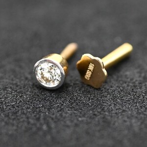 May include: Close-up of two gold and silver stud earrings with a sparkling diamond center. One earring is in focus, showcasing the bezel setting. The other earring's post has a flower-shaped backing with the text "18K S50".