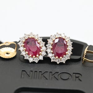 May include: Oval ruby earrings with a diamond halo and gold settings. The image shows the earrings with their backs, and the word "NIKKOR" on a black surface. The earrings are a classic jewellery design.